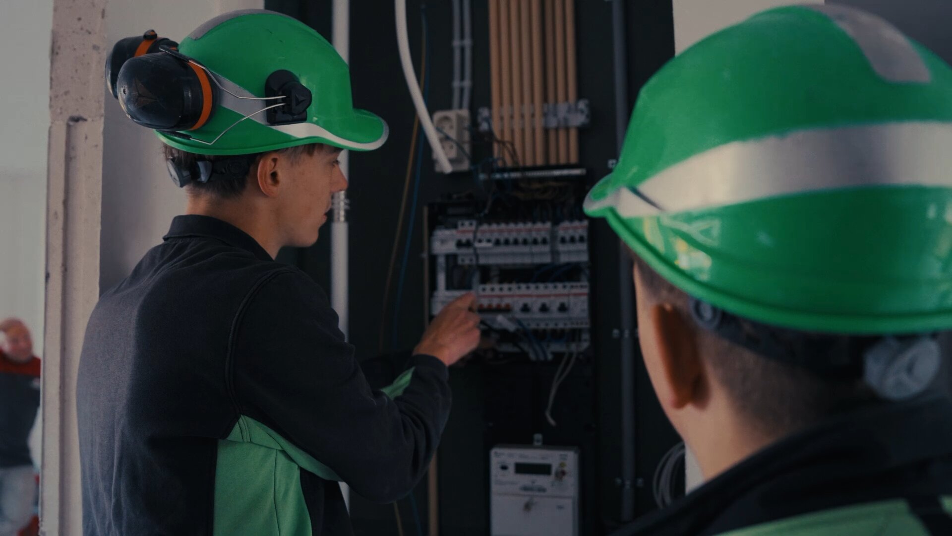 Jonge monteur met groene helm wijst in een groepenkast terwijl een collega meekijkt.