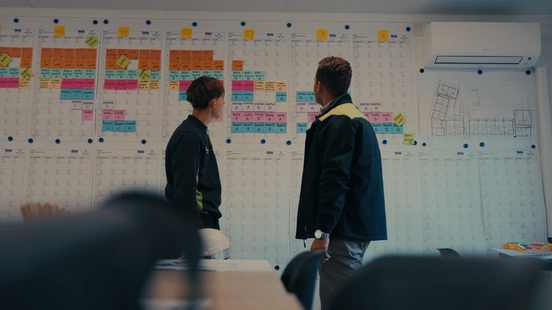 Twee collega's bekijken een bouwplanning vol gekleurde memobriefjes en een plattegrond aan de muur.
