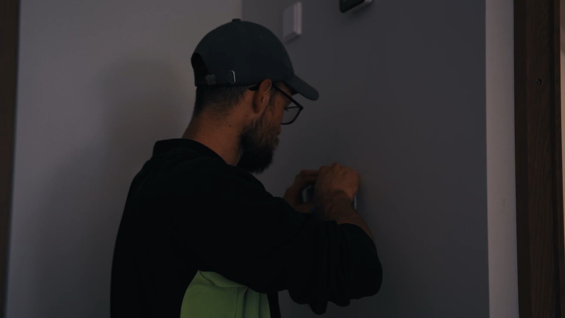 Monteur met pet en bril monteert een onderdeel op de wand in een schemerige ruimte.
