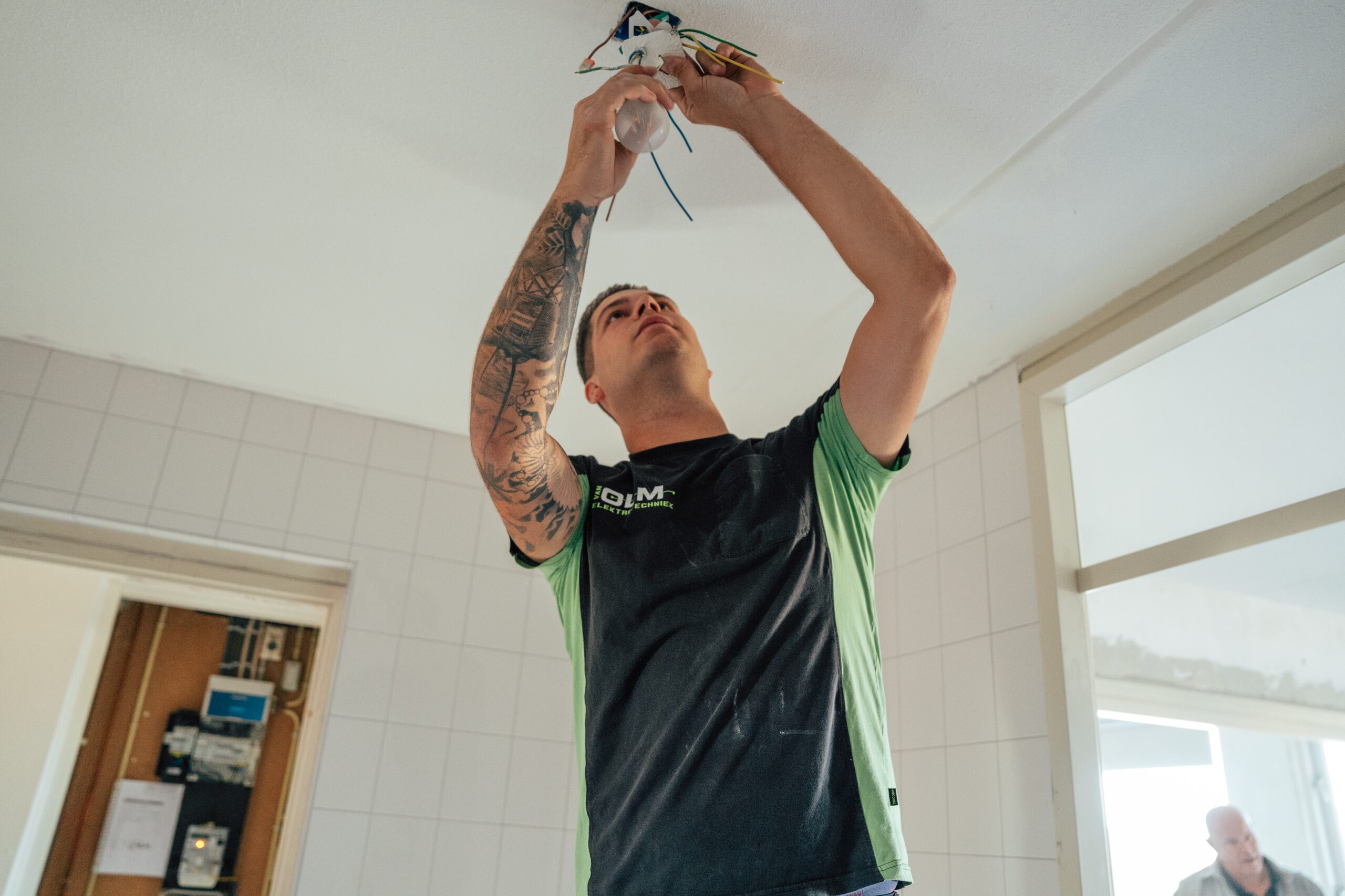 Monteur sluit een lamp aan op het plafond, met de meterkast op de achtergrond.