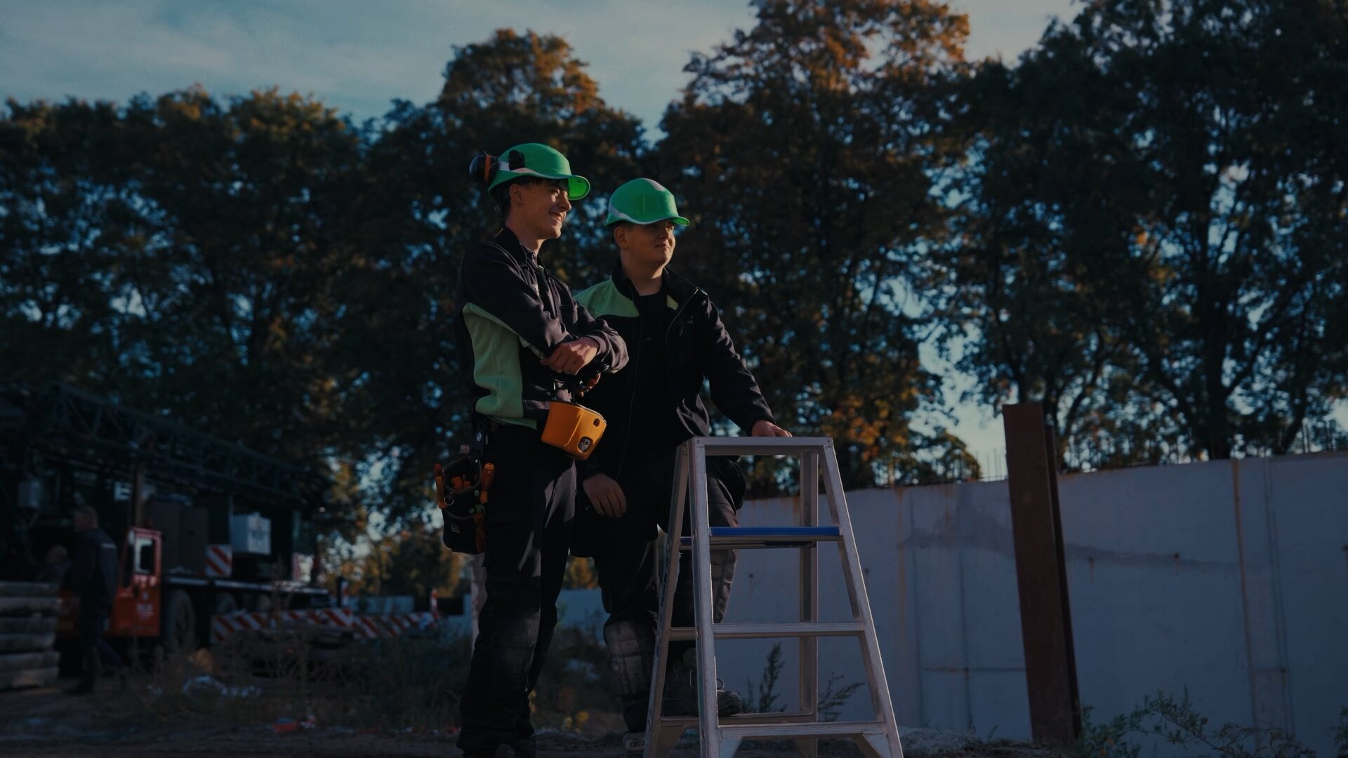 Twee jonge monteurs met groene helm leunen op een trapje op een bouwplaats bij avondlicht.