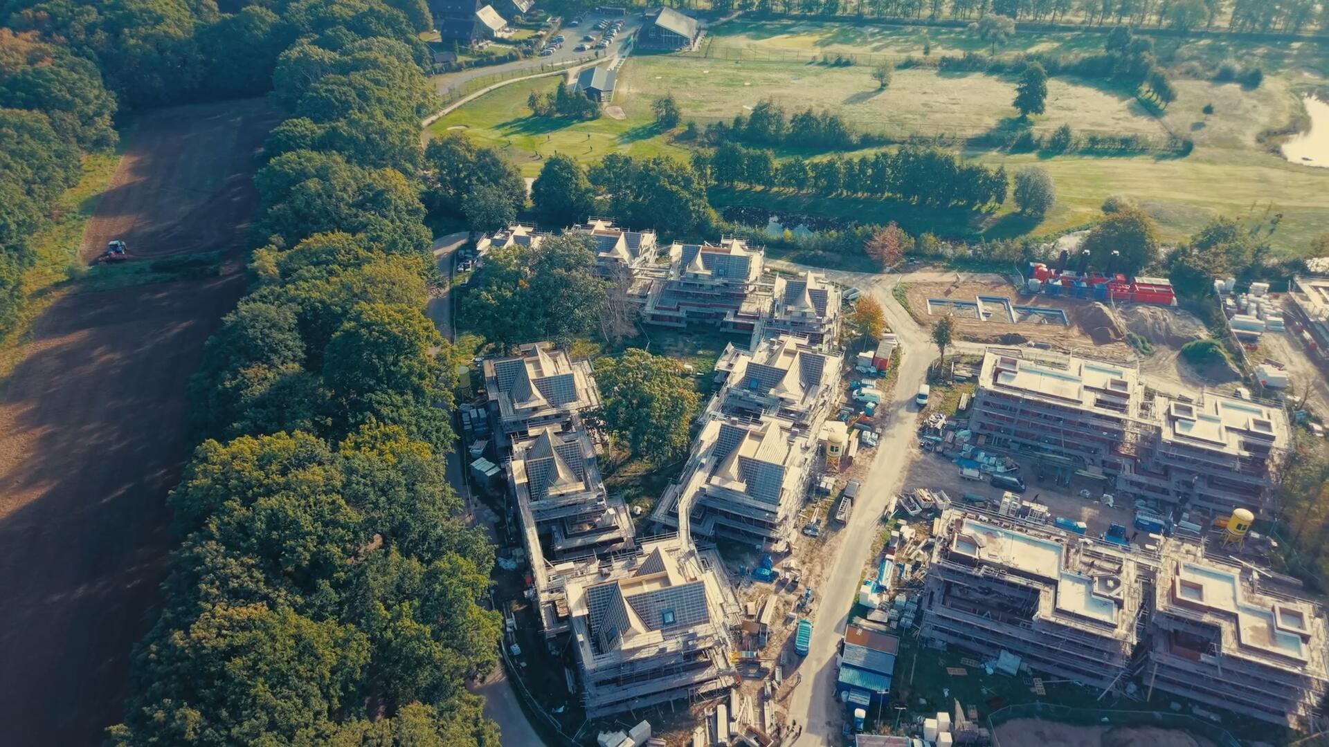 Nieuwbouwproject met tientallen woningen in de steigers, van bovenaf gezien naast een golfbaan.