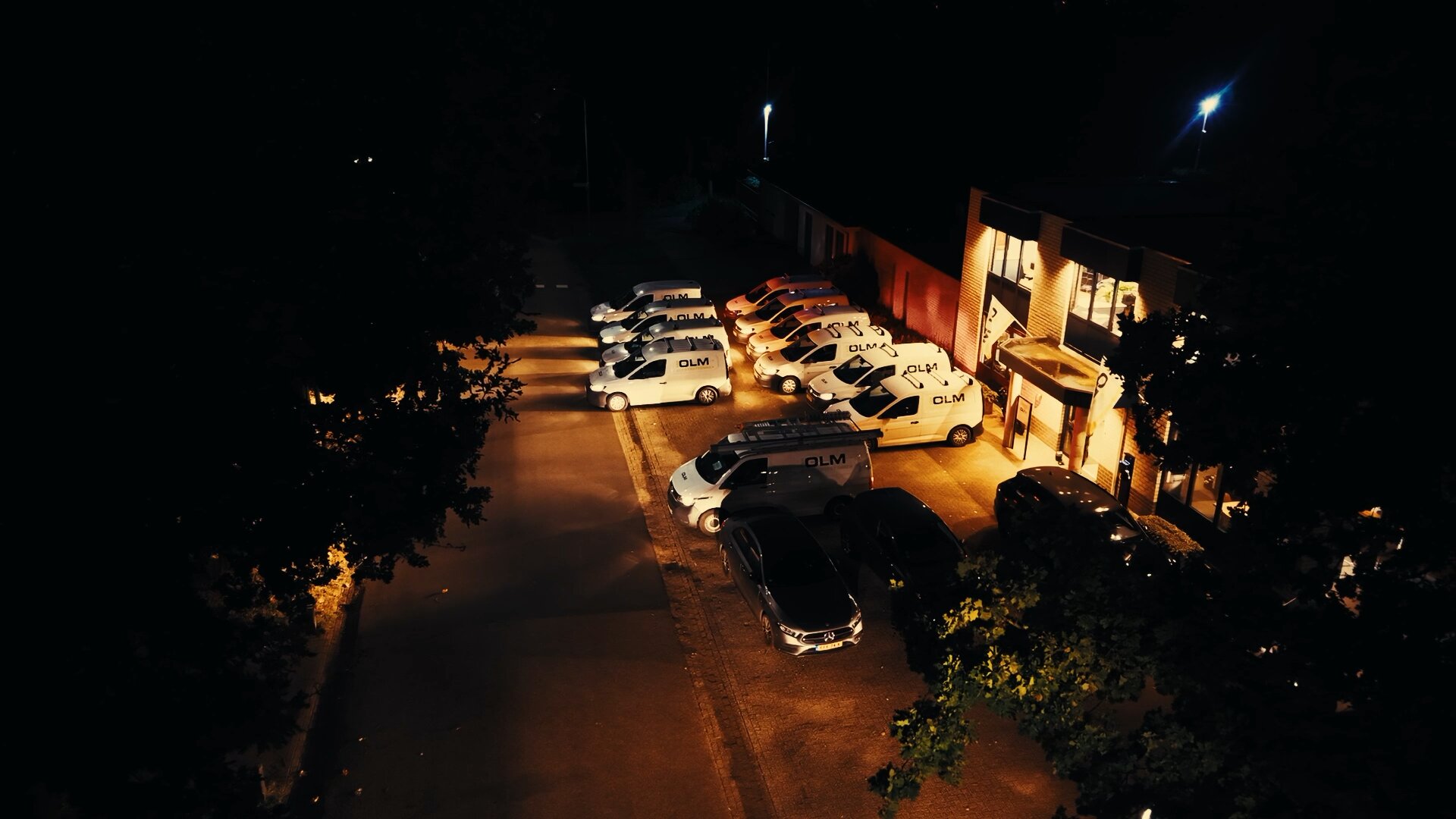 Witte bussen met OLM-logo 's avonds geparkeerd voor het verlichte pand van Van Olm.