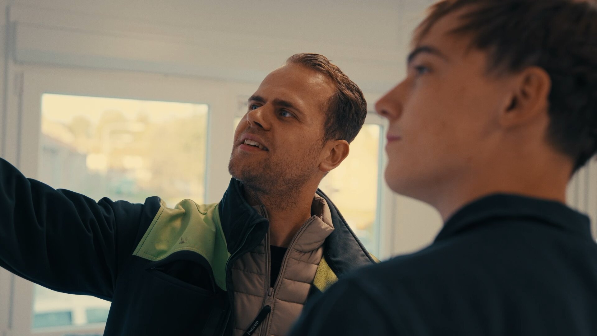 Monteur in werkjas wijst omhoog en legt een jongere collega iets uit in een licht pand.