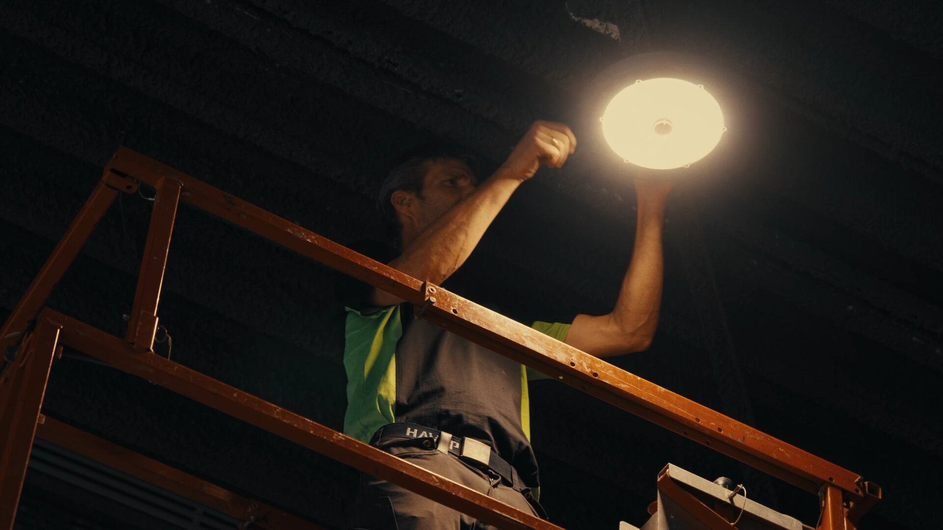 Monteur op een hoogwerker monteert een brandende ronde ledlamp tegen een donker winkelplafond.