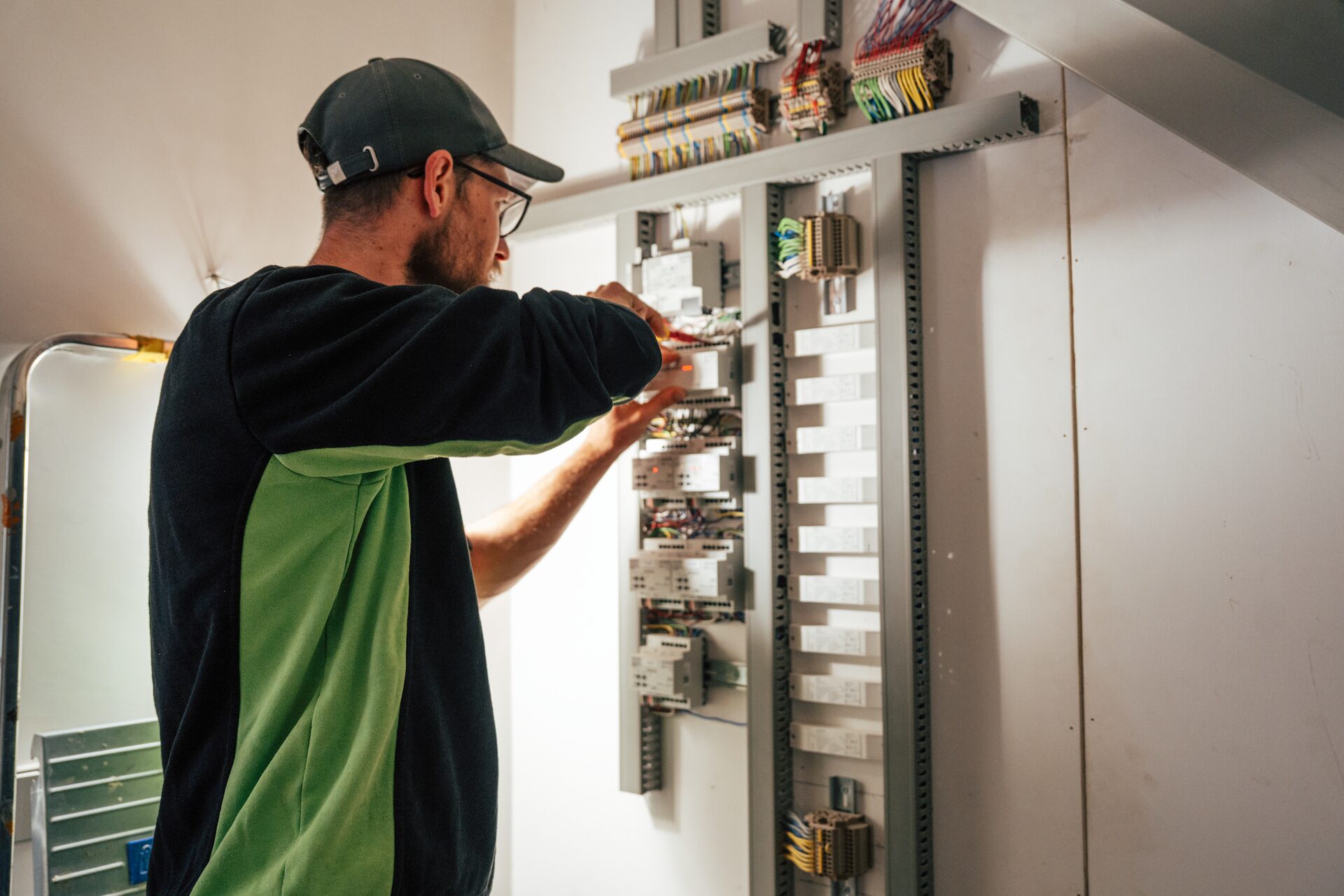 Monteur met pet en bril sluit modules aan in een open verdeelkast vol bedrading.