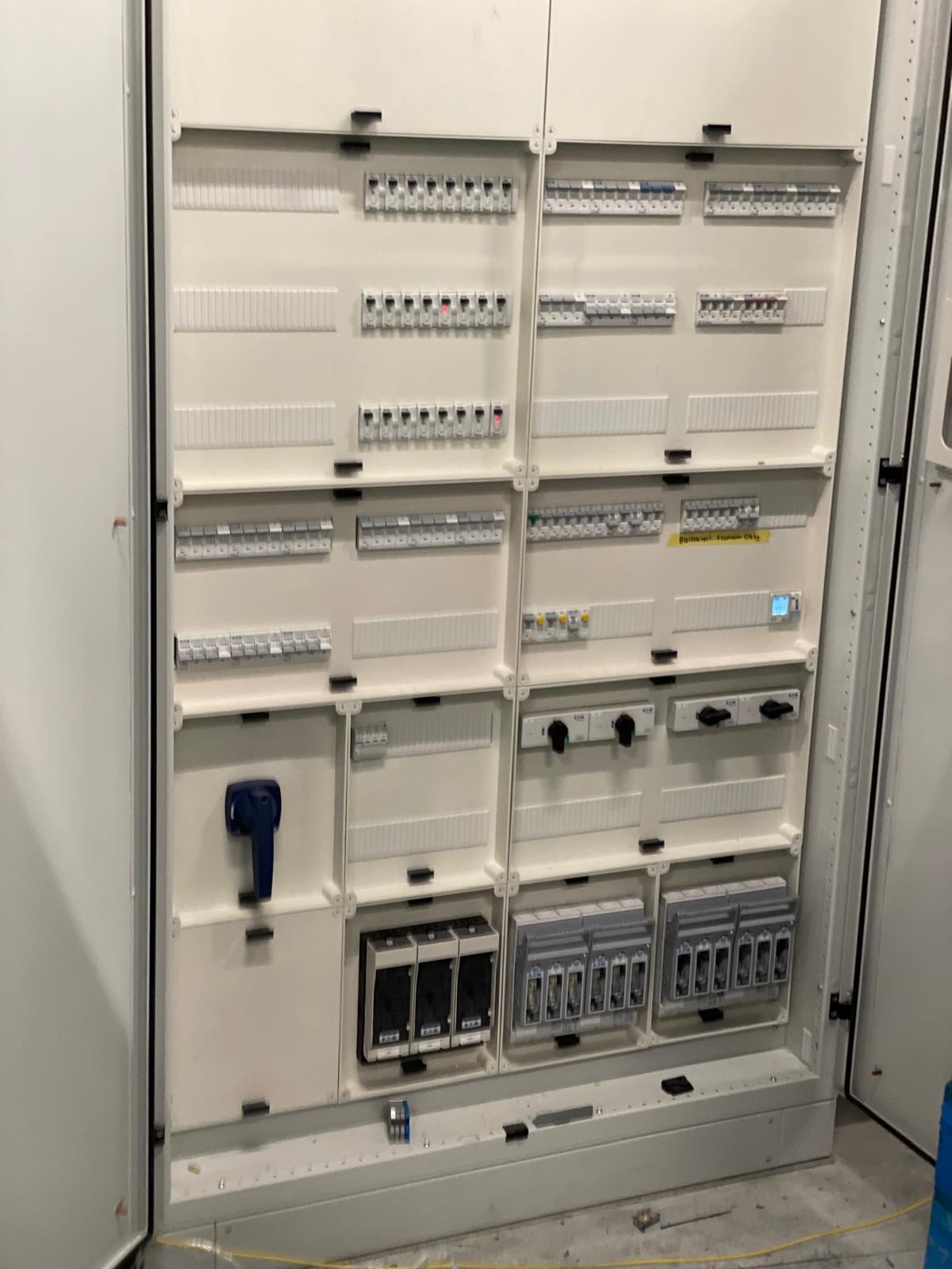 Open hoofdverdeelkast met rijen installatieautomaten, draaischakelaars en een blauwe hoofdschakelaar.