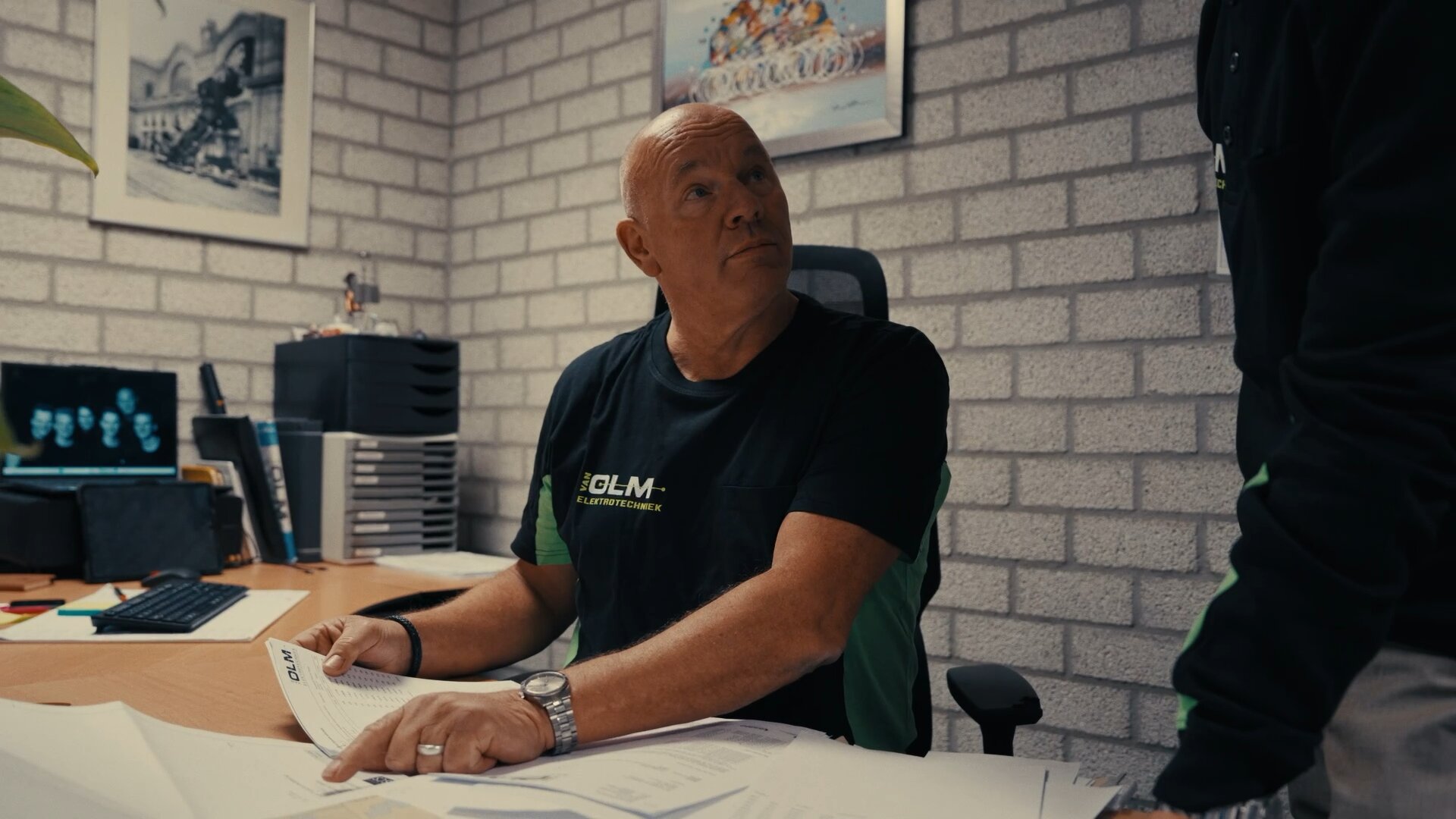 Medewerker van Van Olm Elektrotechniek aan een bureau vol papieren kijkt op naar een collega.