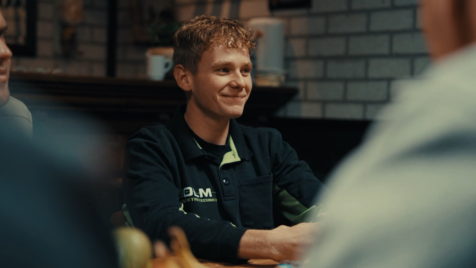 Jonge monteur in Van Olm-werkkleding zit glimlachend aan tafel tussen collega's.