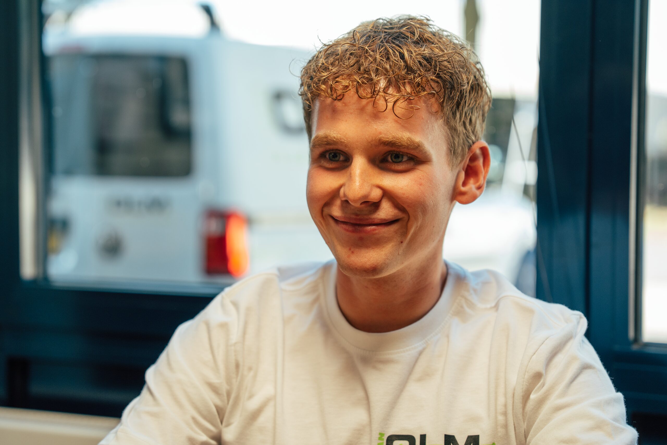 Jonge collega in een wit Van Olm-shirt lacht aan tafel, met een bedrijfsbus buiten.