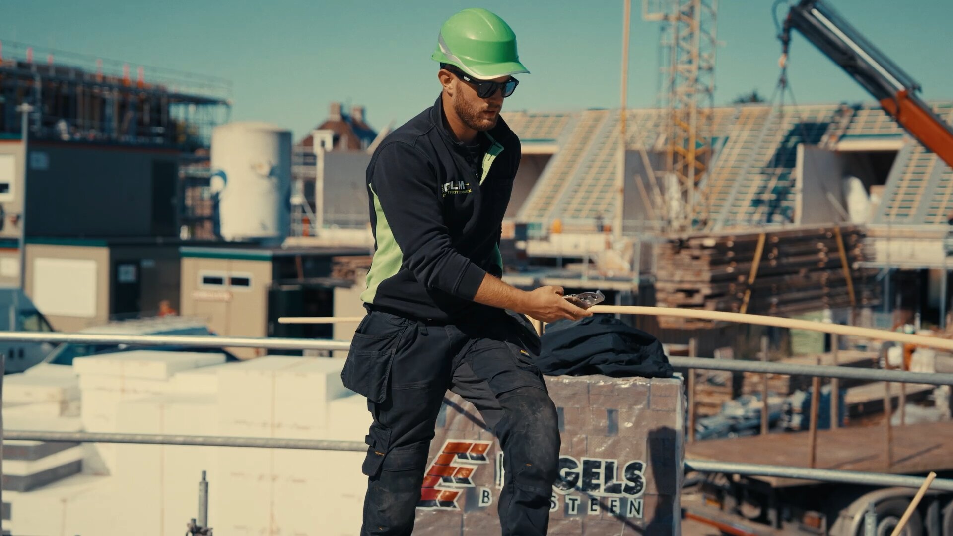 Monteur met groene helm en zonnebril knipt een installatiebuis op maat, met een bouwkraan erachter.