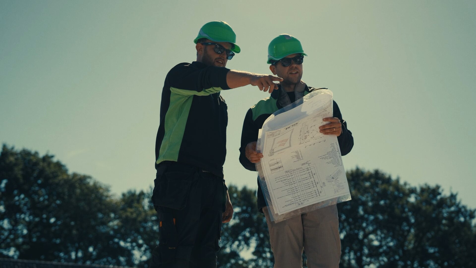 Monteur wijst iets aan op de bouw terwijl een collega de installatietekening vasthoudt.