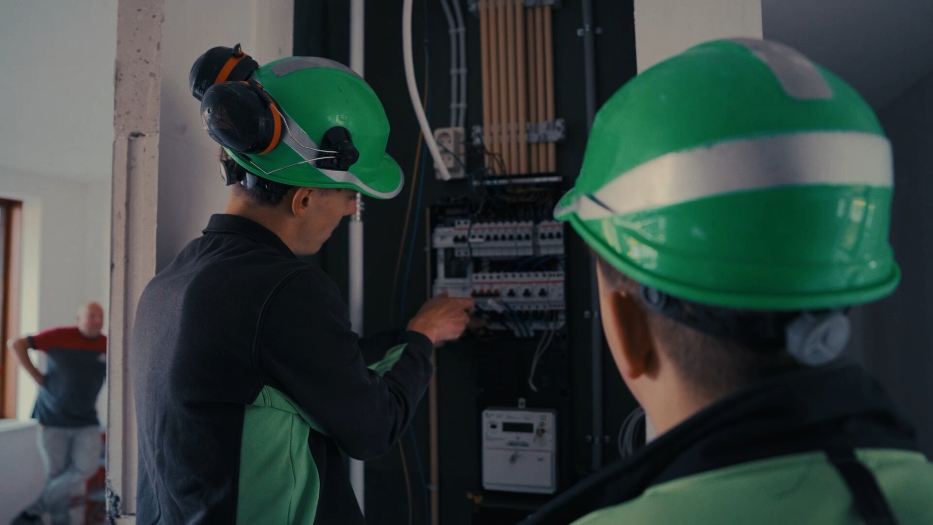 Twee monteurs met groene helmen bij een meterkast in aanbouw, een van hen wijst naar de groepenkast.