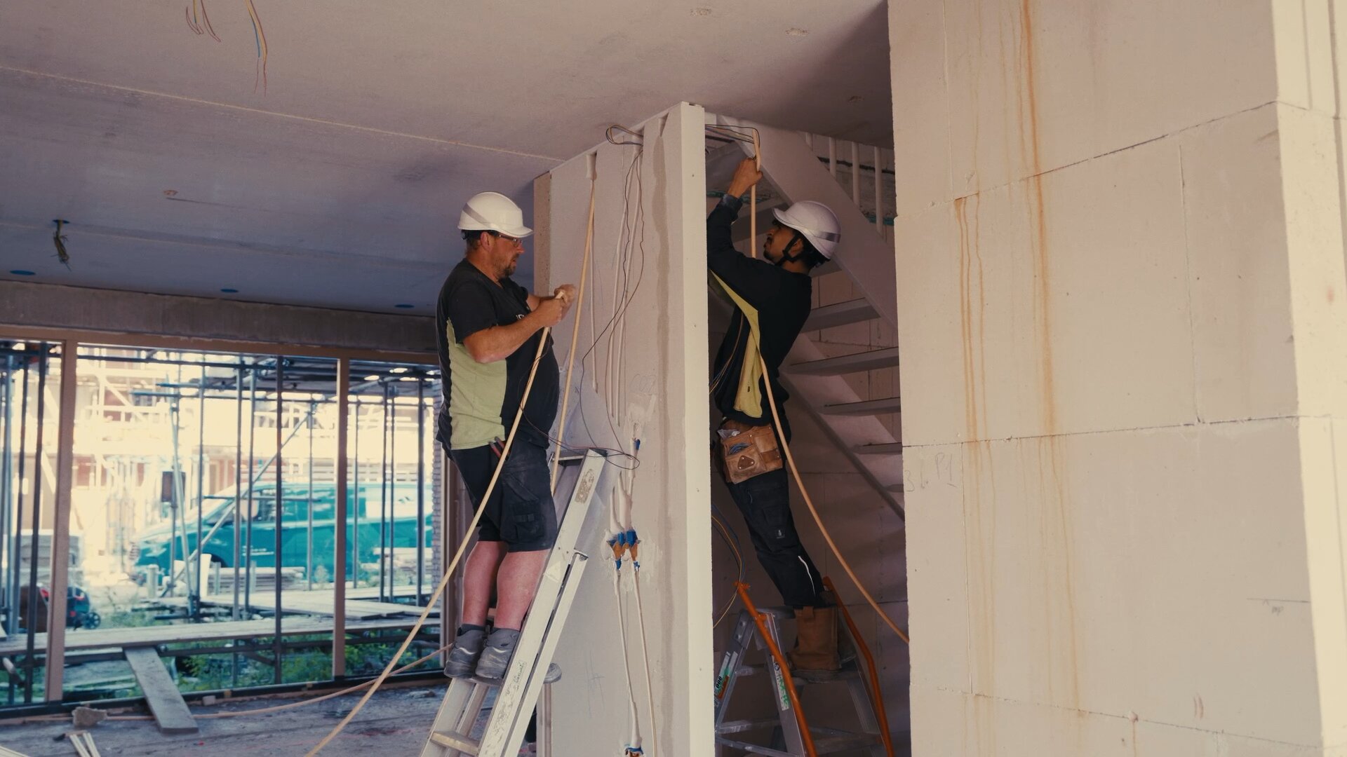 Twee monteurs met witte helmen trekken op trappen kabels door een woning in aanbouw.